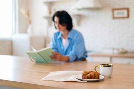 Selective focus on fresh croissants and cup with coffee on wooden table against blurred background with woman reading book. Tasty breakfast in kitchen, spending free time at cozy home.の写真素材