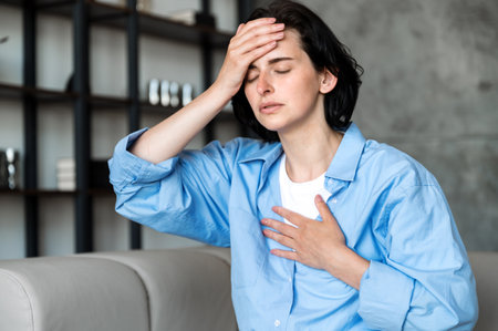 Female having heart, asthma or panic attack. Difficulties with breathe, feeling severe pain. Woman sitting on sofa with closed eyes at home and holding hands on chest and forehead.の写真素材