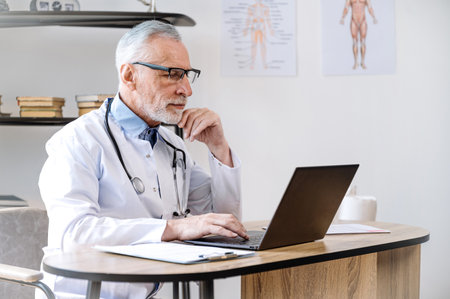 qualified middle aged doctor in glasses and medical coat sitting in clinic cabinet with laptop and searching patient record online while get ready for appointment. health care conceptの写真素材