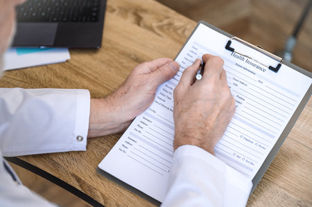 High angle view of family doctor holding clipboard, filling out patient medical information, working...