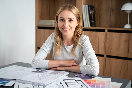 Portrait of smiling young interior designer at home working place. Pantone swatches and house architecture construction plans on table. Choosing best colors for renovatingの写真素材