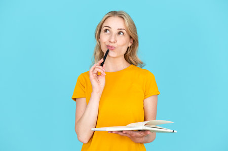 Thoughtful woman holding and keeping pen near chin. Standing pensively against blue background. Thinking about future, consider plan, having dream or idea. Writing thoughts on paperの写真素材