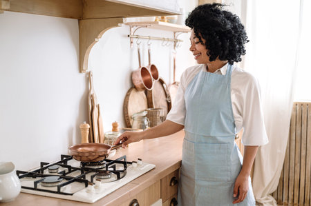 smiling afro american woman in apron put frying pan on gas stove and cooking dinner at home cozy kitchen. cheerful woman prepare helthy food for lunchの写真素材