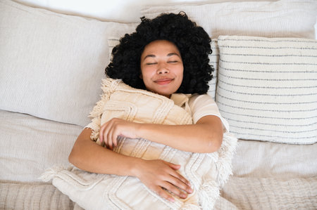 top view of smiling african american woman sleeping in soft bed and hugging pillow. dreamy female with closed eyes resting and imagination in cozy bedroom.の写真素材