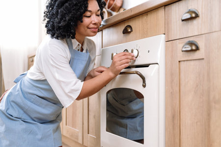 Happy african american housewife in blue apron choosing program for baking food in electric oven. Built-in appliance on wooden cabinet in scandinavian style kitchen.の写真素材