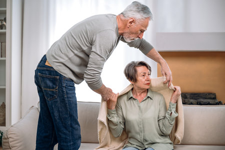 Mature man covered his elderly wife with plaid, while she sitting on couch upset. Concept of supporting, giving psychological empathy and understanding in marriageの写真素材