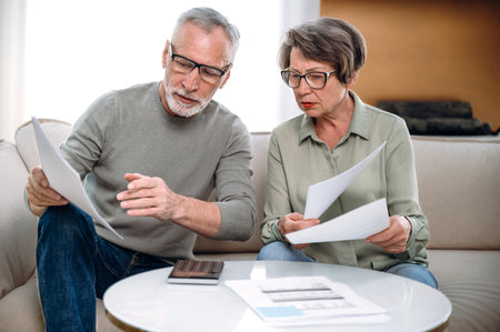 mature couple fill tax form, holding current expense document in hands, while sitting on couch at home. wife and husband receive utility bill, checking banking account scoreの写真素材