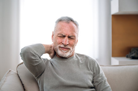 middle aged man feeling discomfort and ache, touching and massaging neck with hand while sitting on sofa at home. tired mature male suffering from nerve compression.の写真素材