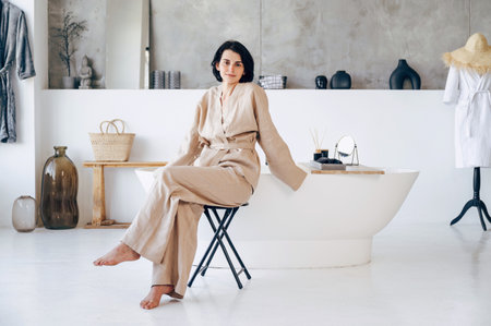 Full length view of calm woman sit on chair against white classic freestanding tub, looking at camera, spending morning at home. Bathroom with loft style interior design. Slow living conceptの写真素材