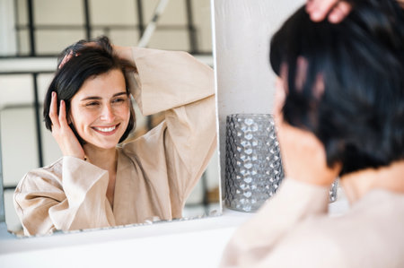 Reflection in mirror of smiling woman touching head and feel enjoyed of healthy hair after washing with professional shampoo. Haircare for dry, damaged, treatment, wellness concept.の写真素材