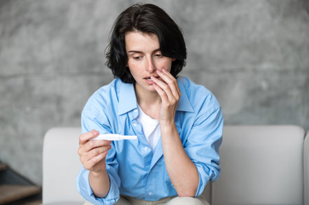 Sad young woman looking at pregnancy test with positive result, feeling confused and think about future while sitting on sofa. Unplanned pregnant conceptの写真素材