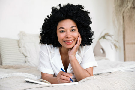 Smiling afro american woman relaxing on bed and writing to-do list in planner. Paper book with ideas and inspiration thoughts. Weekend rest at home. Female student make homework, using diary journalの写真素材