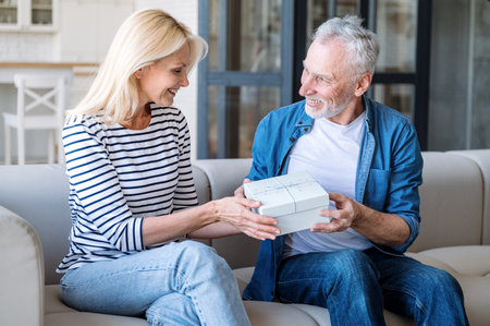 Mature couple exchanging gifts, expressing love. Spouses celebrating special occasion at home, spending romantic weekend together. Man congratulating woman. Making joy to partnerの写真素材