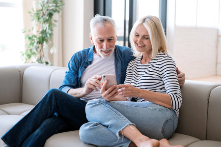Mature spouse using smartphone together, sitting on cozy sofa at home. Smiling man and woman having...