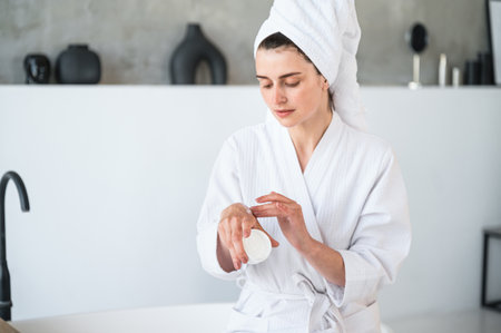 Young woman in white bath robe and towel on head applying moisturizer cream on hand in domestic bathroom. Female using hydration lotion for body after shower. Home spa procedure conceptの写真素材