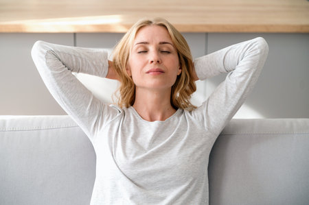 Portrait of calm woman with closed eyes holding hands behind her head, resting at cozy light home. Female enjoying weekend in living room. Recreation, mindfulness and mental health conceptsの写真素材