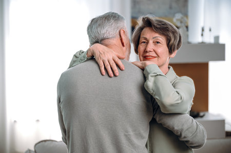 elderly man standing and hugging her wife in living room at home. loving couple resting and spending time together. woman and male support each other in retired old ageの写真素材
