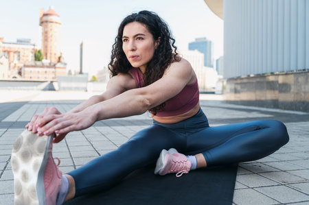 Calm athletic woman sitting on fitness mat and stretching muscles on legs against blurred city...