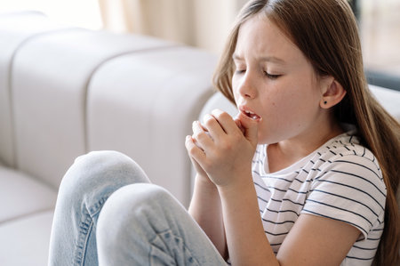 Junior girl sitting on sofa, feeling bad and suffering from dry cough. Kid with respiratory infection symptom or bronchitis staying at home. Healthcare conceptの写真素材