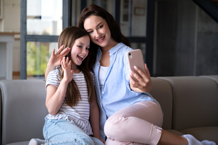 Happy smiling mother and child daughter waving hands, looking at webcam using smartphone for video call. Woman and kid having fun, greeting family online through mobile appの写真素材