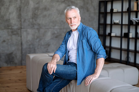 Mature businessman in casual clothes sitting on couch in living room and thinking about work and future plans. Pensive male taking break from work at home, resting and looking awayの写真素材