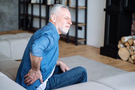 Senior man feeling pain, spasm, backache. Mature male sitting on sofa and have cramps in lower back. Suffering from chronic illness, problem with standing up. Lumbar osteochondrosis, inflammationの写真素材