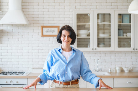 Smiling young woman in casual clothes standing near cooking table on kitchen background and looking at camera, make content for food blog. Female housewife start culinary masterclassの写真素材