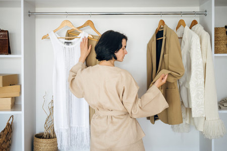 Back view of young woman in beige home suit standing near wardrobe with garments and select outfit...