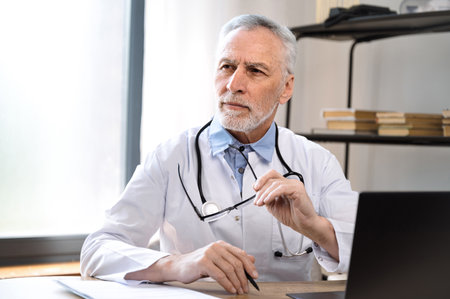 thoughtful serious mature male doctor in white uniform sitting in hospital cabinet, looking in distance and pondering about diagnosis. pensive physician make decision in clinicの写真素材