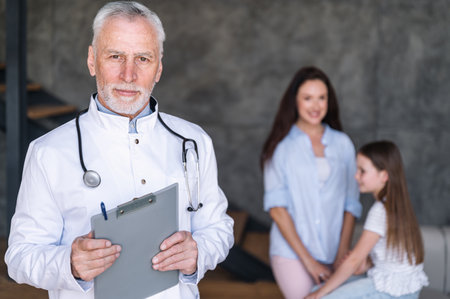 Medical insurance, healthcare concept. Portrait of assured pediatrician with stethoscope and clipboard looking at camera standing on blurred background with patients. Mother and daughter visit clinicの写真素材