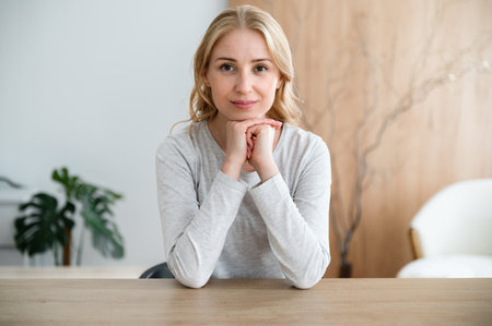 Calm female sitting behind work desk at home office looking at camera, smiling nice. Confident woman work online with clients, recording video for internet blog, provide remotely psychotherapy sessionの写真素材