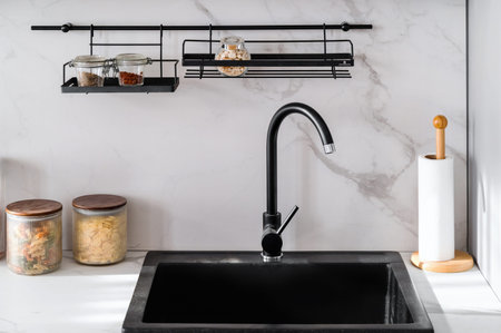 Close up view of black kitchen sink and water tap in modern, minimalist style interior. White marble countertop with food in glass jars. Light ceramic wall with hanging metal shelves against faucet.の写真素材