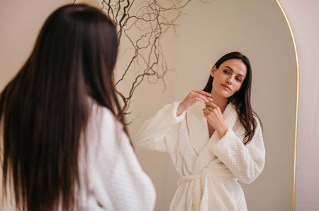 Woman in white bathrobe standing in bathroom after shower, looking at mirror and checking hair damage. Concepts of haircare treatment, beauty and morning routineの写真素材