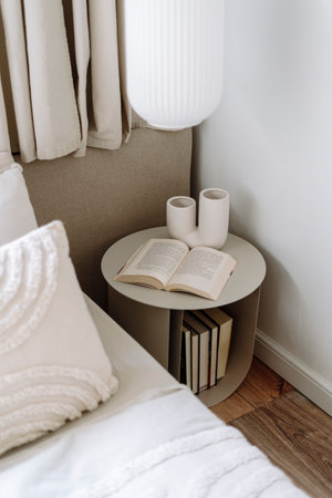 Detail in bedroom with scandinavian interior, high angle view on open book and ceramic vase on round nightstand table near bed with pillows and blanket. Modern home decor in stylish apartment.の写真素材