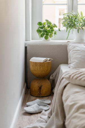 Detail of interior in cozy bedroom, comfort bed with bedding linen and pillows, wooden night side table with book, houseplant on window sill and home slippers on floor. Decor element in apartmentの写真素材