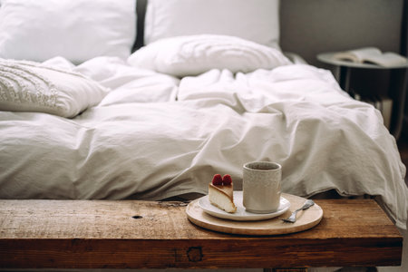 Selective focus on bamboo board with ceramic cup with hot beverage and plate with piece of cake on wooden bench near comfortable bed with linen bedding in cozy bedroom in apartmentの写真素材