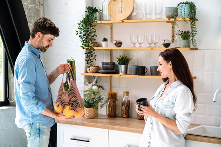 Young man holding groceries in mesh bag standing near woman in modern kitchen at home. Doing household chores together. Healthy diet on weekend.の写真素材