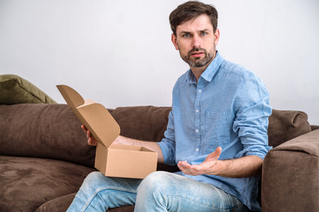 Confused young man getting poor quality goods from online shop. Male sitting on couch, opening box...