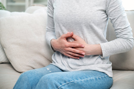 cropped shot of woman with stomach ache feeling discomfort, hold hands on belly and sitting on couch, digestive system problem. sick young female suffering from menstrual painの写真素材