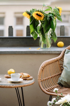 Selective focus on round table with cup of coffee on top and croissant. Rattan chair on summer terrace close to sunflowers in glass vase. Place for rest on balcony. Patio in a buildingの写真素材