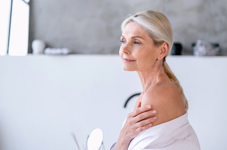 Middle aged 50s woman applying moisturizing body lotion on naked shoulder after morning shower. Advertising of bodycare home spa procedures, anti age treatment, sun protection, beauty products conceptの写真素材