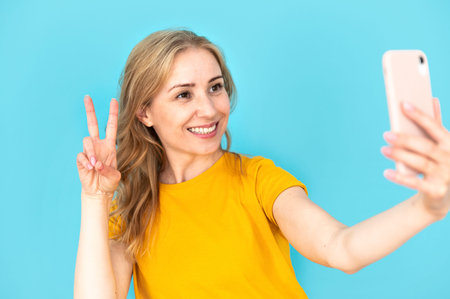 Selfie pose. Happy smiling woman holding smartphone and showing v sign gesture. Taking selfportrait on blue background. Mobile phone photographyの写真素材