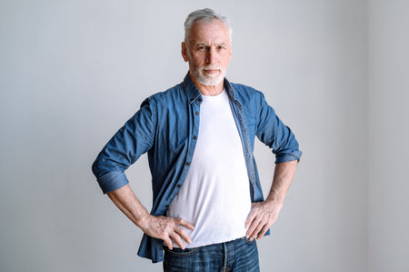 Confident middle aged man wearing denim shirt standing on white background, keeping hands on waist...