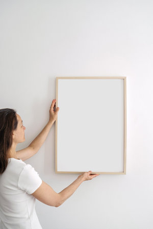 Profile side view of woman in white t-shirt hanging poster in living room. Female holding wooden...
