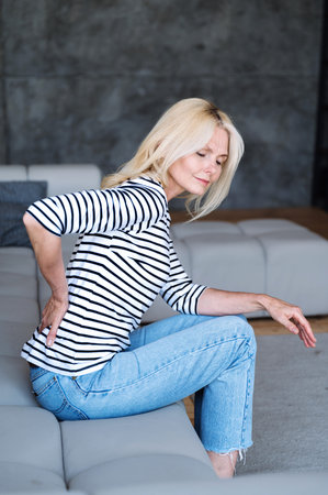 Vertical shot of middle aged woman feeling pain, spasm, cramps in lower back sitting on comfortable...