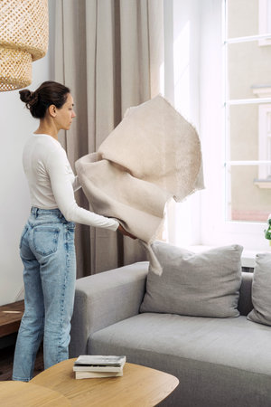 Side view of young woman in casual clothes unfolds a soft blanket over couch. Female holding light...