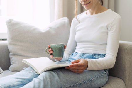 Close up view of female hand holding ceramic cup with coffee. Woman sitting on comfortable couch, enjoying hot beverage, reading notes in notebook. Rest and relax at cozy home on weekend.の写真素材
