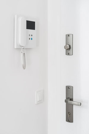 Closeup on intercom system hanging on white wall close to closed front door with metal handle and...