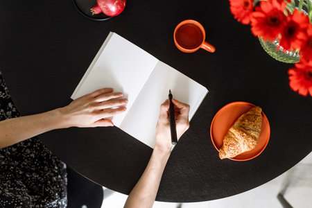High angle view of female hands with pen over diary journal. Woman take notes to paper notebook, working, studying at home, write records to empty planner sit behind desk with morning coffee croissantの写真素材