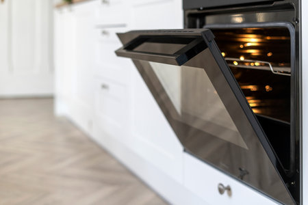 Selective focus on opened glass door with handle of modern electric oven with turning on light in kitchen. Built-in domestic appliances for baking pastry food.の写真素材